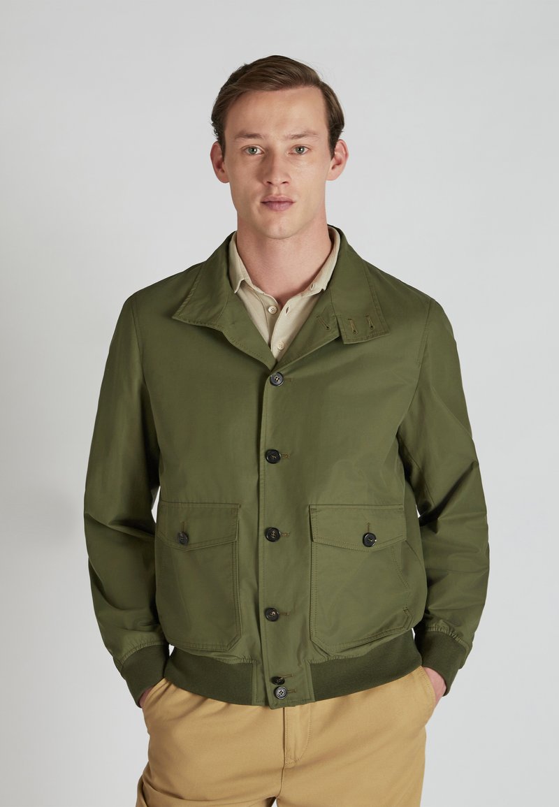 Green lightweight jacket with a pointed collar, button closure, two front pockets, and ribbed cuffs, worn over a beige shirt.