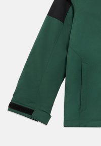 Casaco impermeável verde com detalhes pretos nos ombros. Apresenta um acabamento texturizado, punhos ajustáveis com velcro e um bolso lateral.