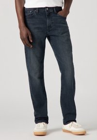 Mörkblå denimjeans med rak bendesign, fem fickor och knäppning, tillsammans med vita sneakers med gummisulor.