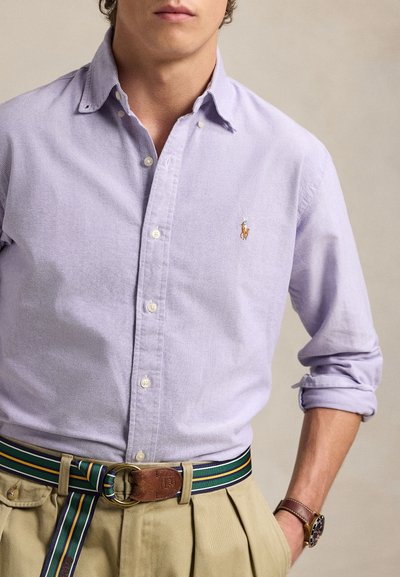 Mężczyzna w jasnofioletowej koszuli na guziki z wyhaftowanym logo, beżowych spodniach, pasku w paski i brązowym zegarku na nadgarstku, pozujący z jedną ręką w kieszeni.