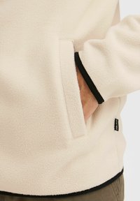 Sudadera de forro polar en color crema con una textura suave, que presenta un bolsillo lateral y ribetes negros en los puños y el dobladillo.