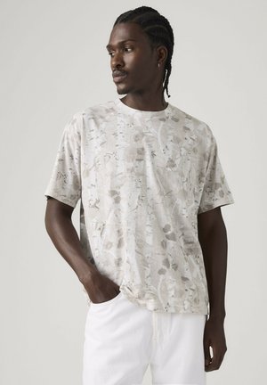 Jeune homme aux cheveux tressés portant un t-shirt beige clair à motifs et un pantalon blanc, regardant vers sa gauche avec une main dans sa poche.