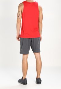 Haut de sport sans manches rouge associé à un short gris foncé. Le matériau semble léger ; le short présente un petit logo près de l'ourlet. Baskets noires.