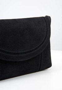 Černá semišová kabelka do ruky s zakřiveným klopou a detailním prošíváním na bílém povrchu a světlém pozadí.