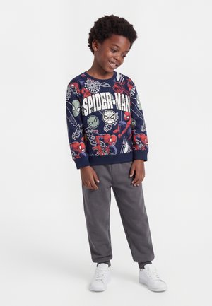 Garçon souriant portant un sweat Spider-Man bleu marine, un pantalon gris et des baskets blanches, debout avec une posture détendue devant un fond uni.