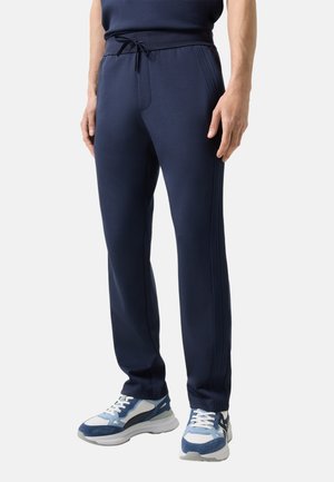 Mężczyzna ubrany w granatowe spodnie dresowe z bocznymi paskami oraz niebiesko-białe buty sportowe, stojący na jednolitym tle.