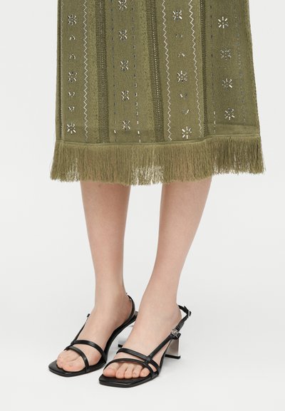 Πόδια με μαύρα σανδάλια με χοντρό τακούνι και λουράκια συνδυασμένα με φούστα λαδί με κρόσσια, διακοσμημένη με κεντητά λουλουδένια και ζιγκ-ζαγκ μοτίβα.