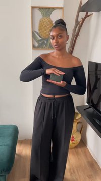 Black off-the-shoulder top with long sleeves, paired with black loose-fit pants; person holds a small rectangular textured green object.