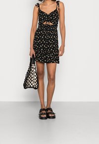 Svart blommig klänning med knytning vid axlarna, utskuren design, rynkad midja och lager-på-lager fåll. Modellen håller en svart och vit vävd väska och bär svarta sandaler.