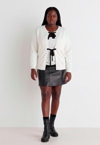 Biely cardigan s detailom mašle vpredu, preložený cez pruhovaný čierno-biely top, skombinovaný s čiernou koženou mini sukňou a členkovými čižmami.