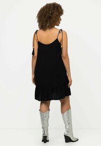 Vestido negro sin mangas con lazos ajustables de borlas, espalda fruncida y dobladillo con volantes. Combinado con botas plateadas brillantes hasta la rodilla.