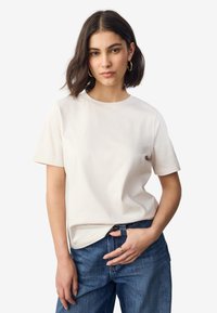 T-shirt beige clair à manches courtes en coton. Il présente un col rond et une coupe décontractée, accompagné d'un jean en denim bleu.