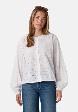 Jeune femme aux cheveux châtain clair porte un chemisier blanc à œillets avec manches ballon et un jean bleu, des lunettes de soleil sur la tête.