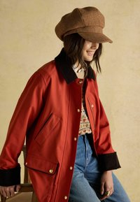 Punane veekindel jope manšetiä�ärega, kuldsete nööpidega, küljetaskudega, kantud mustrilise kampsuni ja teksade peal.