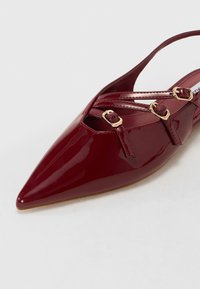 Escarpin en cuir verni bordeaux avec bout pointu, présentant des brides entrecroisées et des boucles dorées pour une touche de détail.