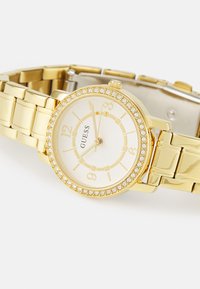 Gold wristwatch with a white face, gold numerals, and a crystal-studded bezel. Features a linked metal band and a polished finish.