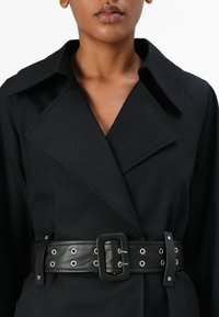 Zwarte trenchcoat met een brede kraag, voorzien van een leren riem. De riem heeft zilveren oogjes en een rechthoekige gesp voor sluiting.