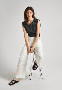 Top sin mangas gris, pantalones blancos con patrones geométricos texturizados, sandalias negras, sentado en un taburete de metal, fondo neutral.
