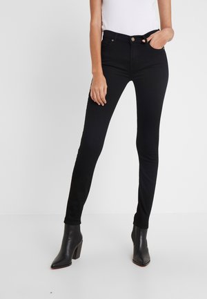 Kvinde iført tætsiddende sorte jeans, hvid ærmeløs top og sorte ankelstøvler med hæl, stående med den ene hånd i lommen.