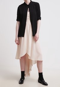 Chemise noire à manches courtes portée sur une robe plissée pêche clair avec un ourlet asymétrique, accompagnée de chaussettes noires et de chaussures en cuir verni.