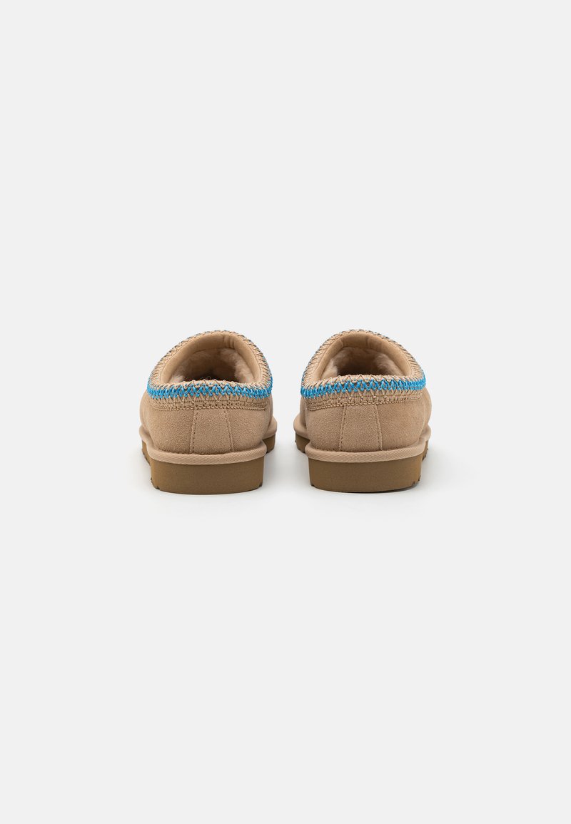 Beige suède pantoffels met een blauwe gestikte accent rond de kraag en een platte, tan rubberen zool. Zacht voering is zichtbaar vanuit de achterkant.