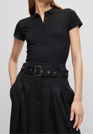 Vrouw draagt een aansluitend zwart poloshirt met korte mouwen en een zwart rokje met hoge taille, voorzien van een brede riem met metalen gesp.