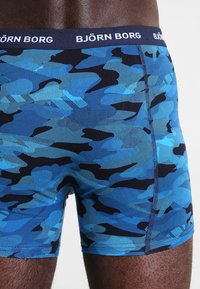Boxers en bleu avec un motif camouflage, dotés d'une ceinture noire portant le logo "BJÖRN BORG". Le tissu a une texture lisse avec des nuances contrastées.