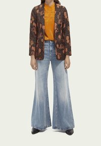 Czarny żakiet w kwiatowy pomarańczowy wzór, pomarańczowa bluzka i jasnoniebieskie dżinsy w fasonie dzwonów. Czarne botki dopełniają stylizację.