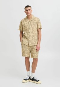 Ensemble en shirt et shorts de couleur beige avec un léger motif floral. Fabriqué dans un tissu léger, assorti à des sneakers noires et des chaussettes blanches.
