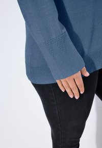 Modrý pletený svetr s texturovanými rukávy, zdobenými lemy a volným střihem, zkombinovaný s černými džíny. Detailní záběr na ruku odpočívající na džínách.