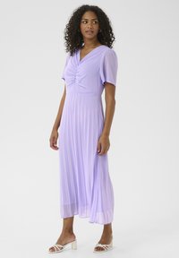 Rochie midi plisată lavandă, cu mâneci scurte, un decolteu plisat și o fustă lejeră. Este purtată cu sandale albe cu toc.