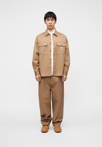 Μπεζ βαμβακερό πουκάμισο-σακάκι με δύο τσέπες στο στήθος, κούμπωμα με κουμπιά στο μπροστινό μέρος και λογότυπο, συνδυασμένο με άνετο μπεζ παντελόνι και καφέ παπούτσια.