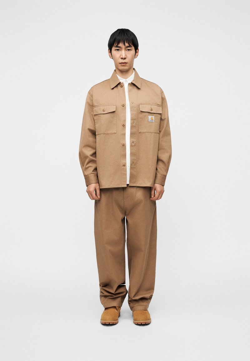 Giacca camicia di cotone color sabbia con due tasche sul petto, chiusura con bottoni e toppa con logo, abbinata a pantaloni larghi color sabbia e calzature marroni.