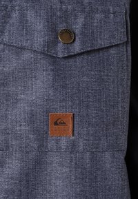 Poche en tissu bleu foncé avec un bouton-pression en métal portant l'étiquette "Quiksilver" et une patch en cuir brun avec un logo représentant une vague et une montagne.