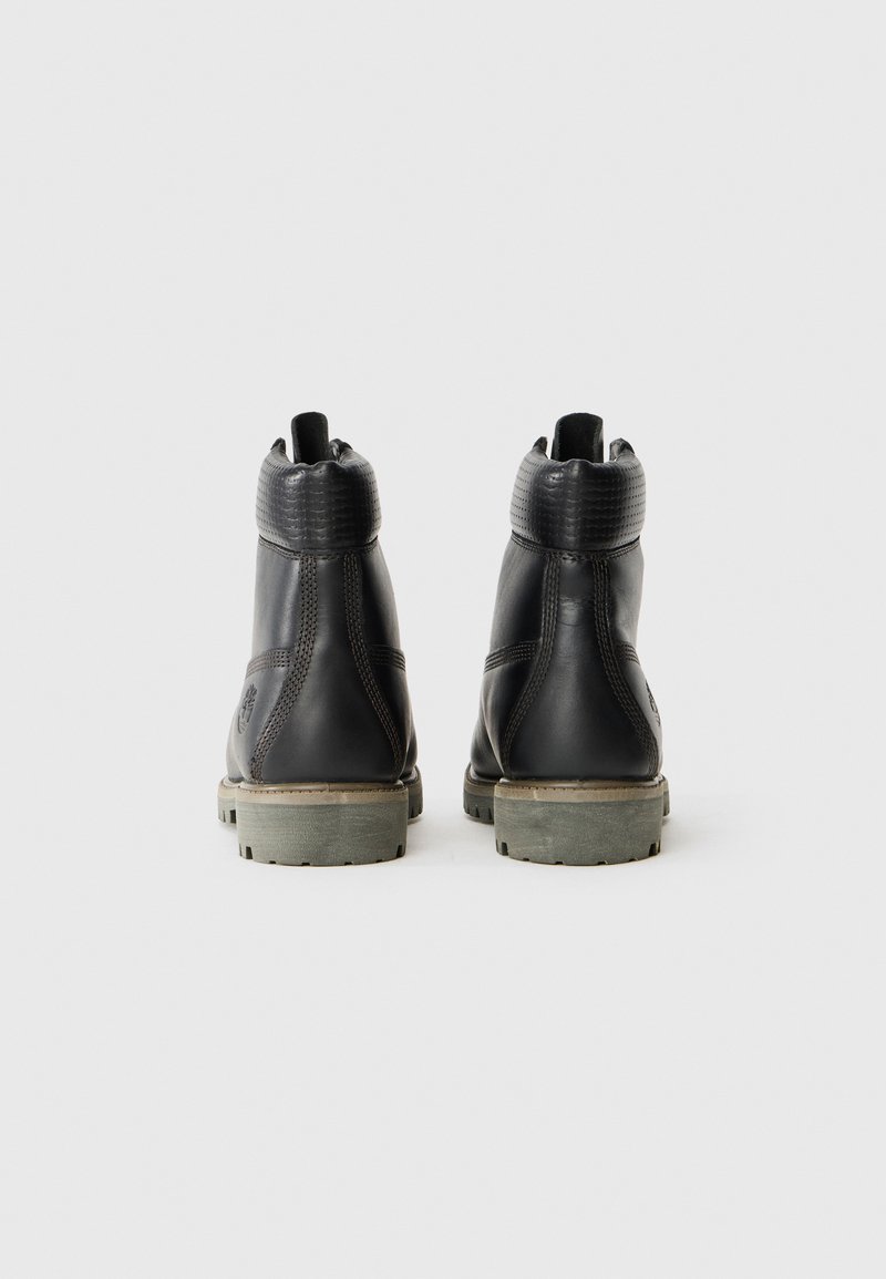 Ein Paar schwarze Lederstiefeletten mit gepolsterten Schaft und tanfarbenen Gummisohlen, von hinten auf einem schlichten weißen Hintergrund betrachtet.