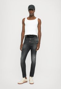 Jeans negros ajustados con ligeras desgastadas, combinados con una camiseta sin mangas blanca y zapatillas beigas. El modelo está usando una gorra negra.