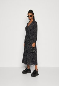 Zwart jurk met stippen, lange mouwen en een strik in de taille, gecombineerd met stevige zwarte platform schoenen en een kleine handtas.