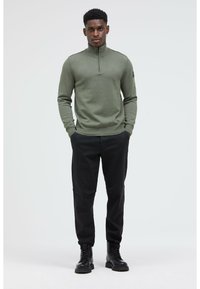 Mann in olivgrünem Quarter-Zip-Pullover, schwarzen Hosen und schwarzen Schnürboots, der mit den Händen in den Taschen vor einem weißen Hintergrund steht.