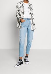 Persona vistiendo jeans de tiro alto color azul claro, camiseta blanca, camisa de cuadro blanco y negro, y zapatillas negras de caña alta, de pie sobre un fondo sencillo.