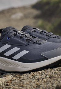 Chaussures de course Adidas gris foncé avec des bandes blanches sur une semelle extérieure texturée, placées sur un chemin de gravier avec un arrière-plan naturel flou.