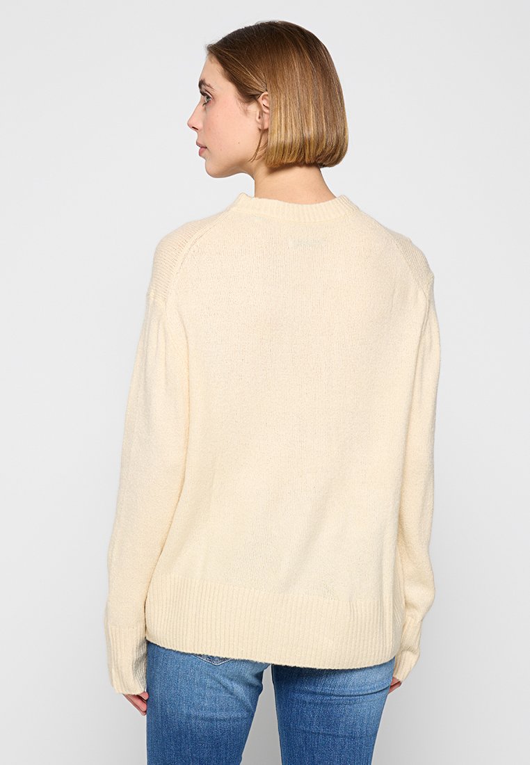 Pull en tricot couleur crème avec une coupe décontractée, des poignets et un ourlet côtelés, et un tissu légèrement texturé, porté sur un jean bleu, vu de dos.