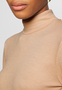 Kerman barkot turtlenecki, joka sisältää tiukasti istuvan muotoilun, sileän tekstuurin ja korkean kaula-aukon. Saumayksityiskohtia on näkyvillä kauluksessa ja hartioilla.
