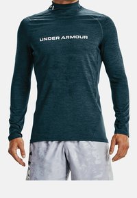 Tmavě modré sportovní tričko s dlouhými rukávy a vysokým límcem, s bílým logem "UNDER ARMOUR" na přední straně a texturovanými detaily látky.