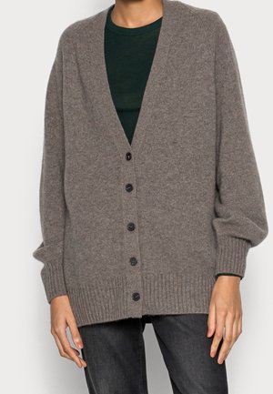Personne portant un cardigan en maille marron ample avec cinq boutons noirs sur une chemise vert foncé et un pantalon sombre, debout devant un fond uni.
