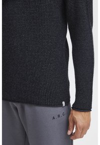 Un pull en tricot noir avec un motif texturé et des manches longues, associé à un pantalon de survêtement gris avec des lettres noires imprimées sur le côté.