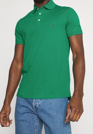 Man die een groen poloshirt met korte mouwen en knopen draagt, gecombineerd met een blauwe spijkerbroek, staand tegen een effen achtergrond.