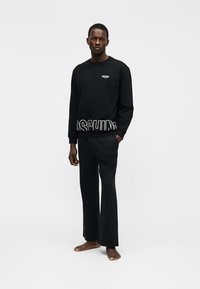 Άνδρας που στέκεται ξυπόλυτος φορώντας μαύρο φούτερ Moschino και μαύρο παντελόνι, μπροστά σε απλό φωτεινό φόντο.