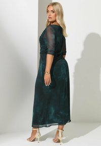 Kvinde med blond hår iført en mørkegrøn, gennemsigtig lang kjole og guld højhælede sandaler, som ser over sin skulder mod en neutral baggrund.