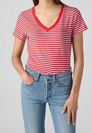 Kobieta w czerwono-białej, pasiastym T-shircie z dekoltem w serek i krótkimi rękawami oraz jasnoniebieskich, wysokich jeansach, stojąca z jedną ręką w kieszeni.
