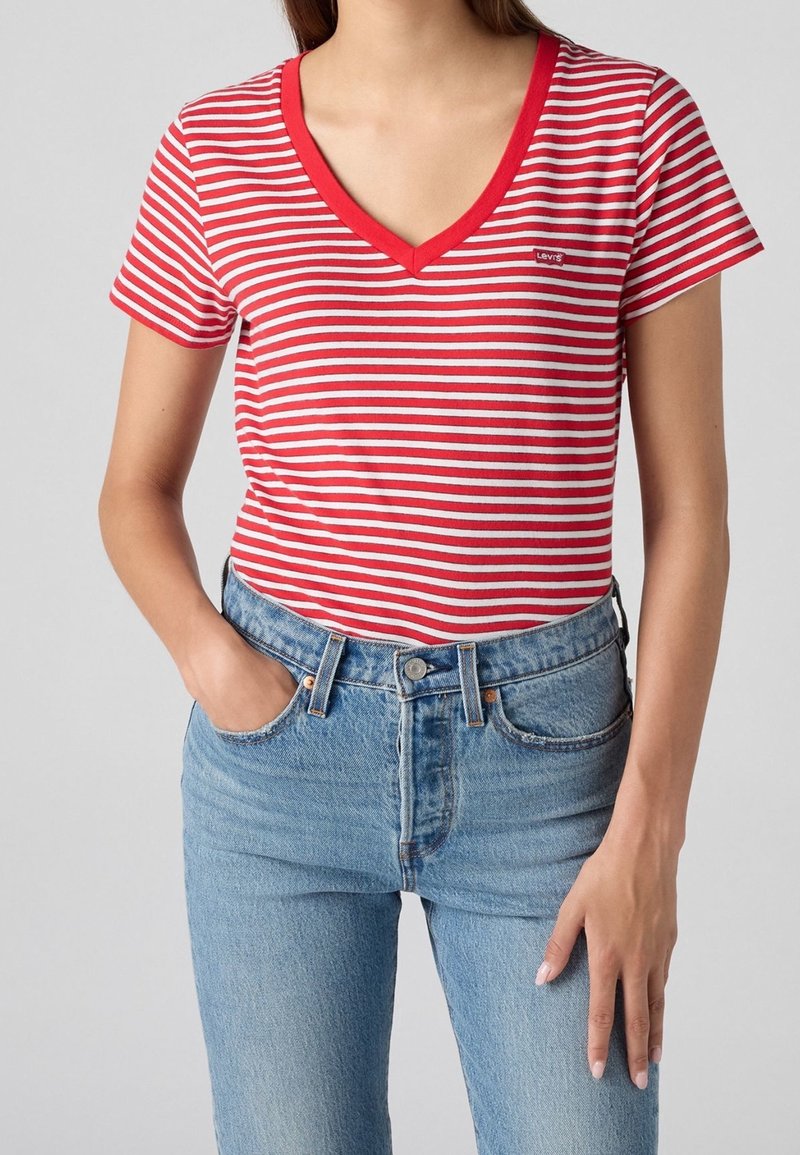 Femme portant un t-shirt à rayures rouges et blanches avec col en V et manches courtes, et un jean bleu clair taille haute, debout, une main dans la poche.